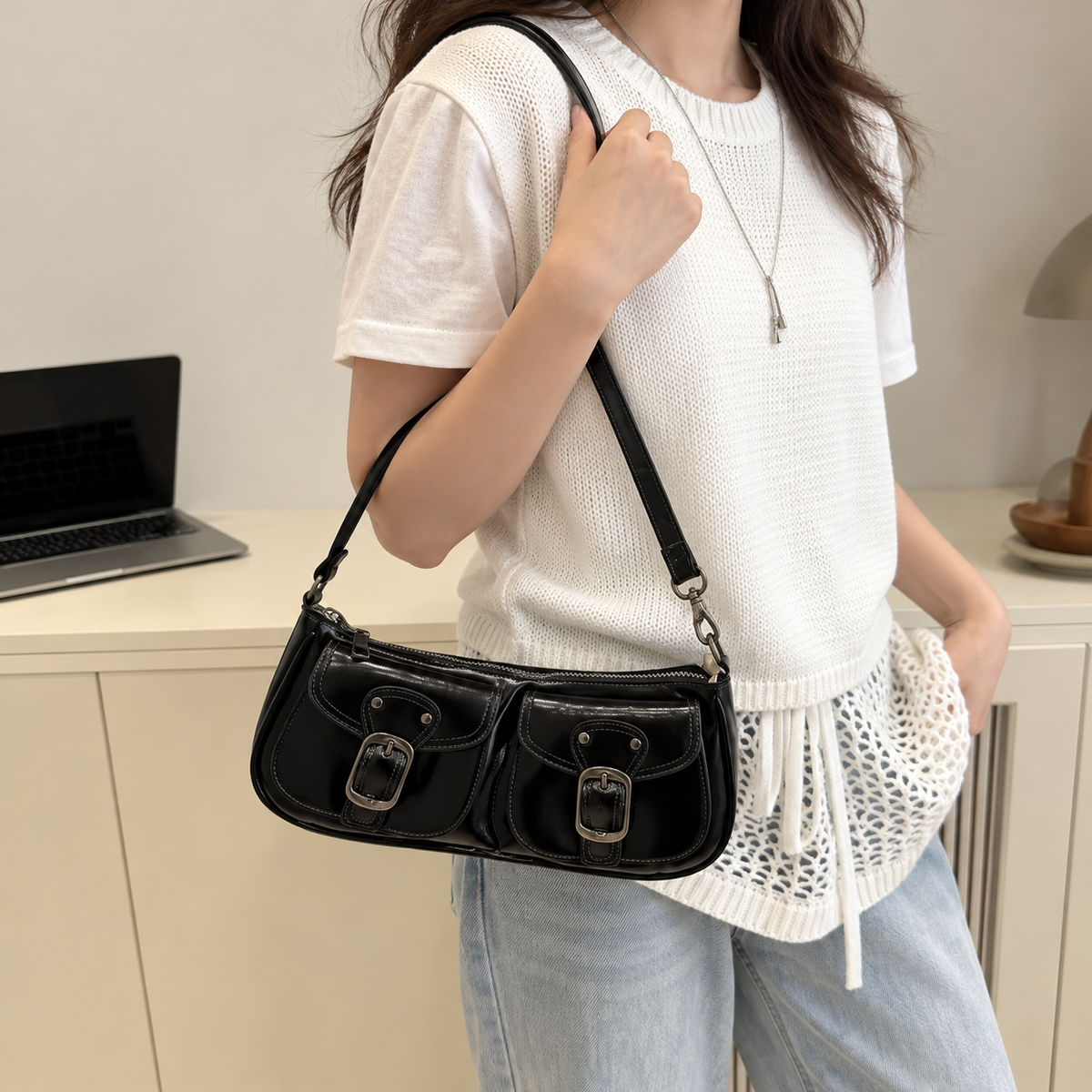 Woman wearing a small black PU leather shoulder bag with twin buckle details beside a white knit top and jeans