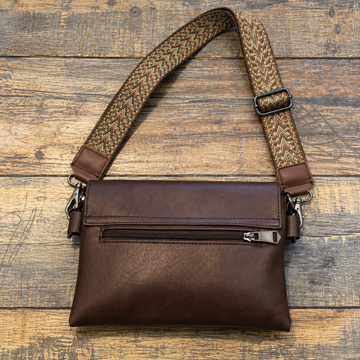 Rear view of a dark brown crossbody bag showing an exterior zipper pocket and an adjustable patterned strap on a wooden surface.