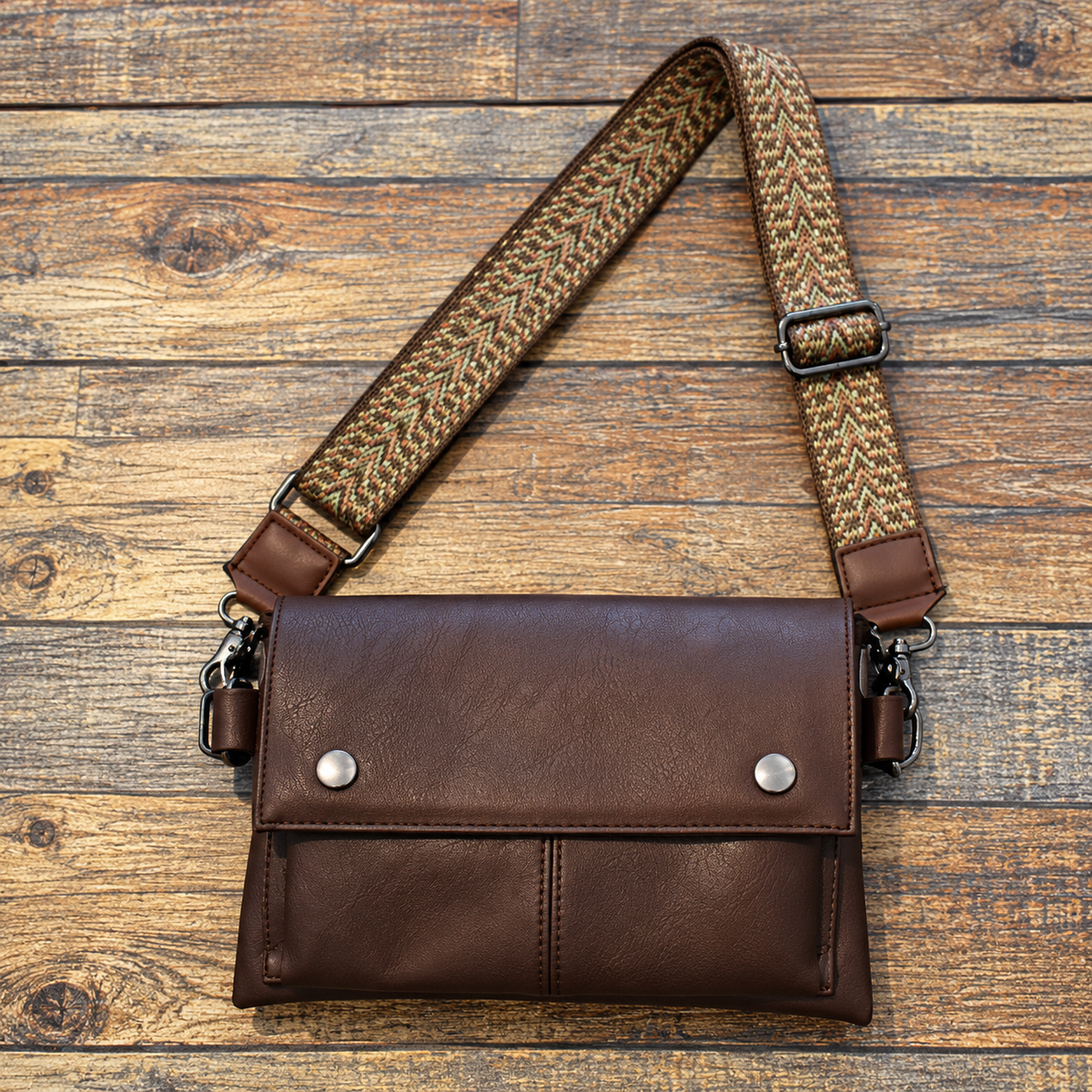 Dark brown PU leather crossbody bag with a front flap, metal details and a patterned shoulder strap on a wooden surface.