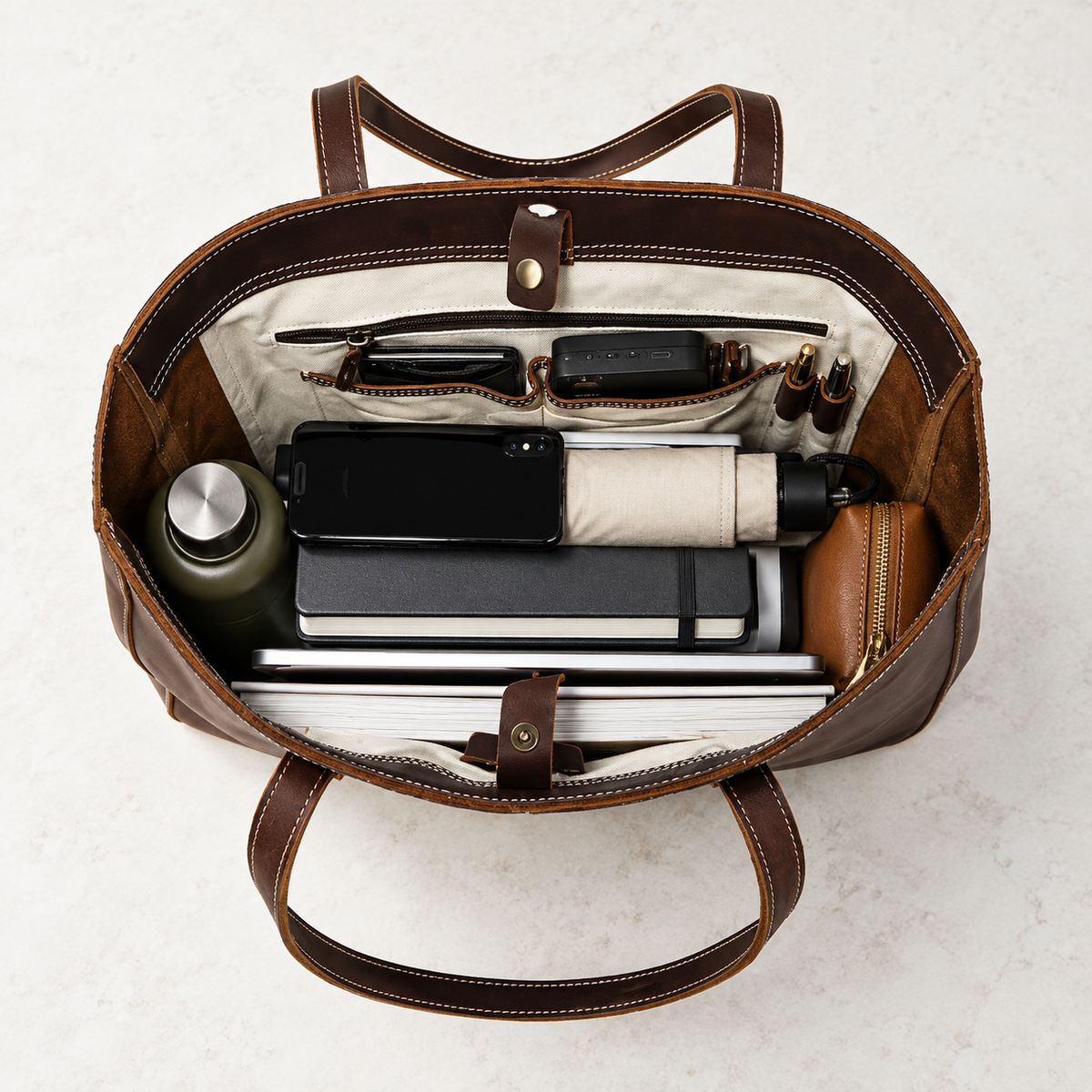 Brown leather tote bag holding a laptop, notebook, phone, umbrella, water bottle and small accessories